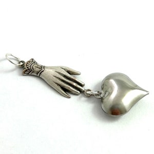 Puede incluir: Un collar con un dije de plata que representa una mano que se acerca a un coraz&oacute;n. La mano est&aacute; detallada con un brazalete y dedos. El coraz&oacute;n es liso y redondeado.