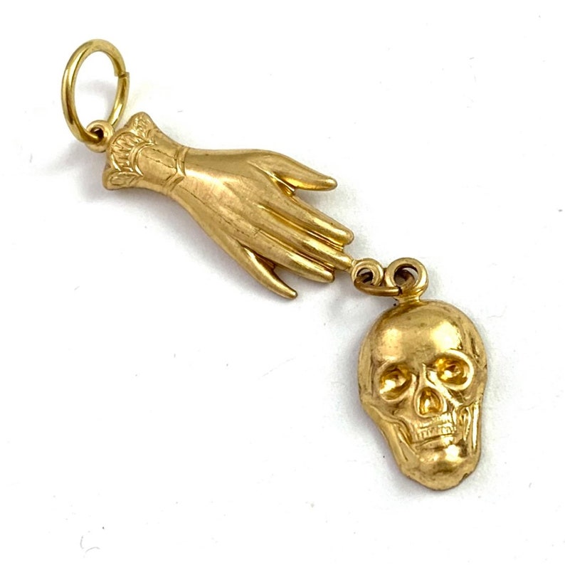 Op de afbeelding: Een gouden bedel met een hand die naar een schedel reikt. De hand is gedetailleerd met een manchet en de schedel heeft een gedetailleerd gezicht.