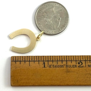 May include: A gold-colored horseshoe pendant with a small loop for a chain. A quarter coin is next to the pendant for size comparison. A wooden ruler shows the pendant is approximately 1 inch wide.