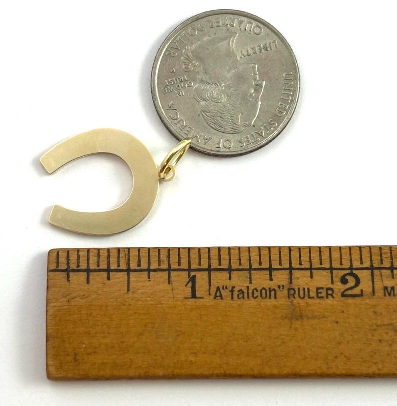 May include: A gold-colored horseshoe pendant with a small loop for a chain. A quarter coin is next to the pendant for size comparison. A wooden ruler shows the pendant is approximately 1 inch wide.
