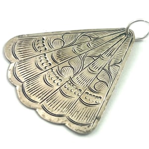 Puede incluir: Colgante plateado con forma de abanico. Presenta intrincados patrones grabados, incluyendo l&iacute;neas curvas y elementos decorativos. Un peque&ntilde;o anillo est&aacute; unido en la parte superior para colgarlo. Los bordes tienen un dise&ntilde;o festoneado.