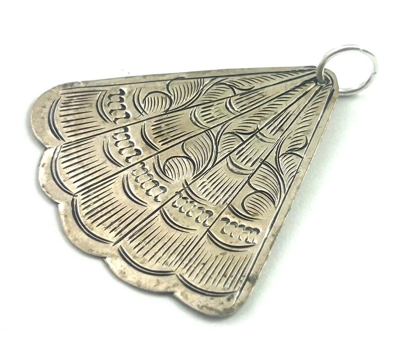 Puede incluir: Colgante plateado con forma de abanico. Presenta intrincados patrones grabados, incluyendo l&iacute;neas curvas y elementos decorativos. Un peque&ntilde;o anillo est&aacute; unido en la parte superior para colgarlo. Los bordes tienen un dise&ntilde;o festoneado.