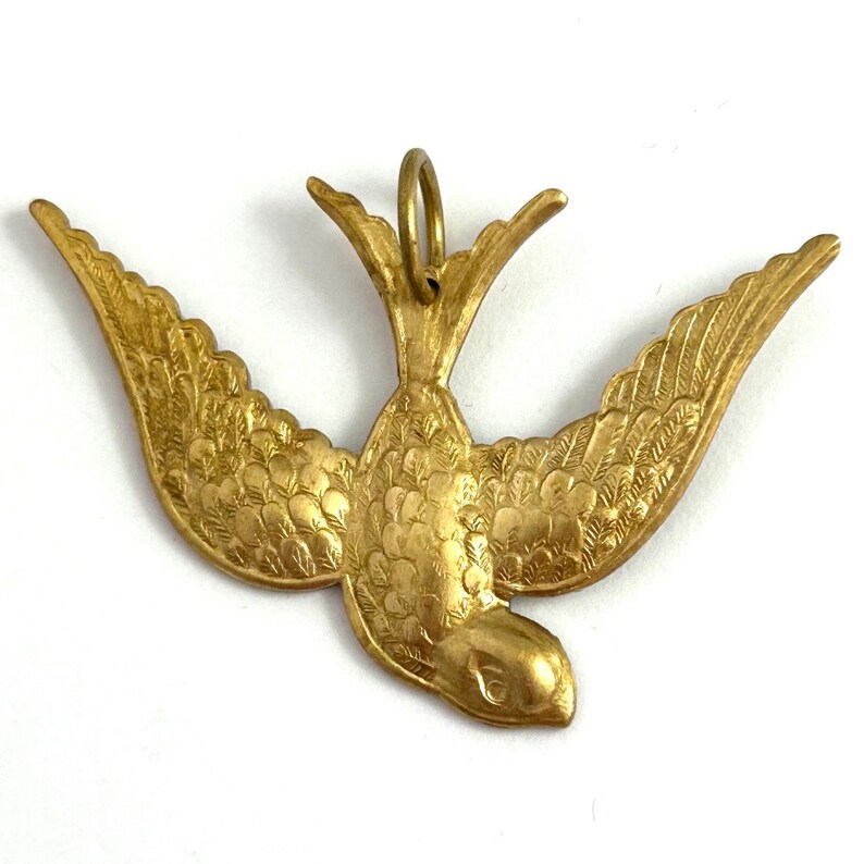 May include: A gold-colored metal pendant in the shape of a stylized bird with its wings spread. The bird has a detailed design with textured feathers.
