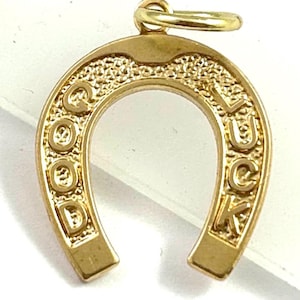 May include: A gold-colored horseshoe charm with the words "GOOD LUCK" embossed on it. The charm has a textured surface and a small loop at the top for attaching to a chain or bracelet. The horseshoe is a classic symbol of good fortune.
