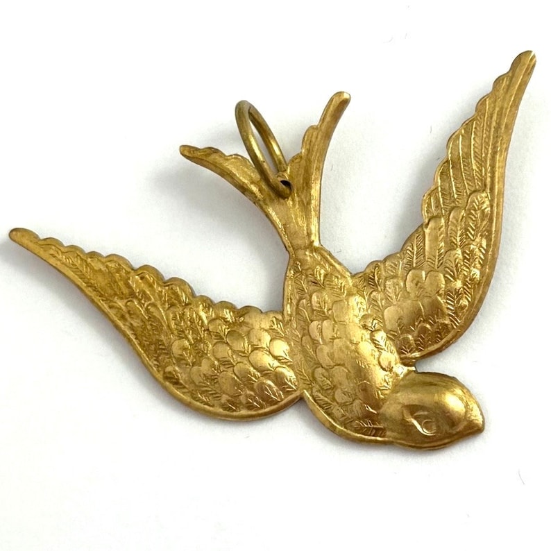 May include: A gold-colored metal pendant in the shape of a stylized bird with its wings spread. The bird has a detailed design with feathers and a small loop at the top for hanging.