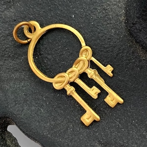 Op de afbeelding: Een gouden bedel met een cirkel waaraan een tros van vier sleutels hangt. De sleutels zijn met een decoratief knoopje aan elkaar gebonden.
