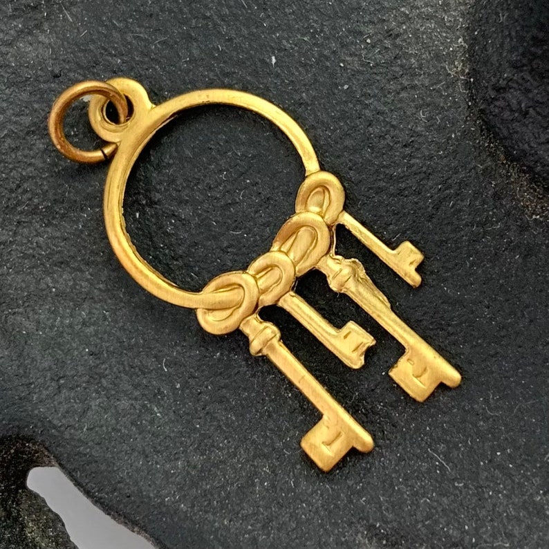 Op de afbeelding: Een gouden bedel met een cirkel waaraan een tros van vier sleutels hangt. De sleutels zijn met een decoratief knoopje aan elkaar gebonden.