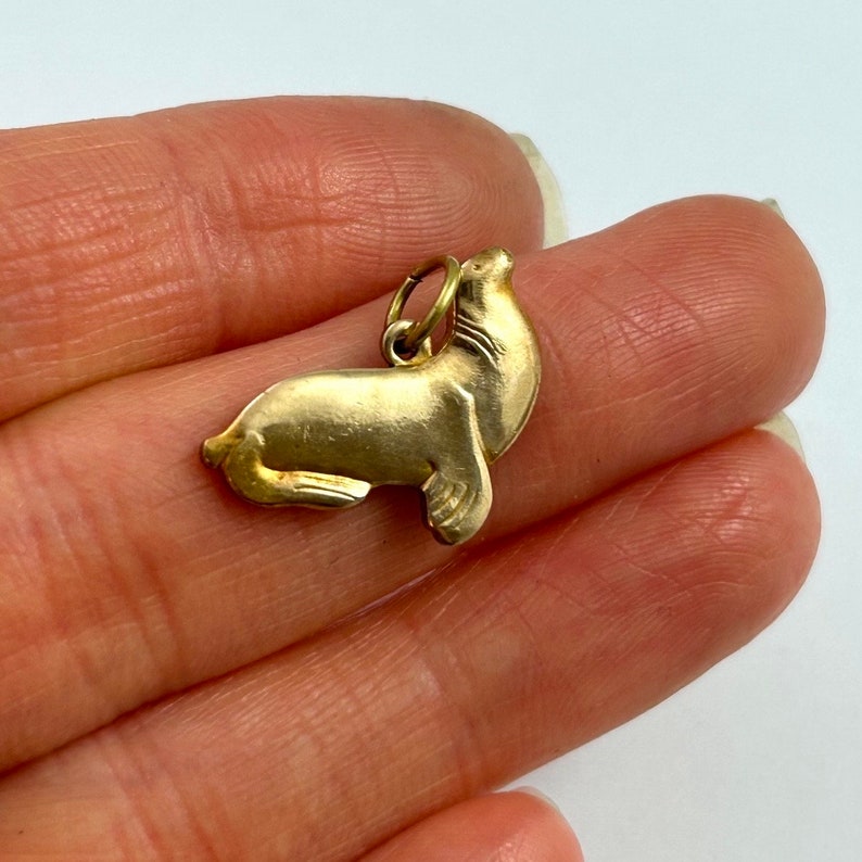 May include: A gold-colored charm in the shape of a seal. The seal is facing upwards with its head tilted back. The charm is detailed with a textured surface.