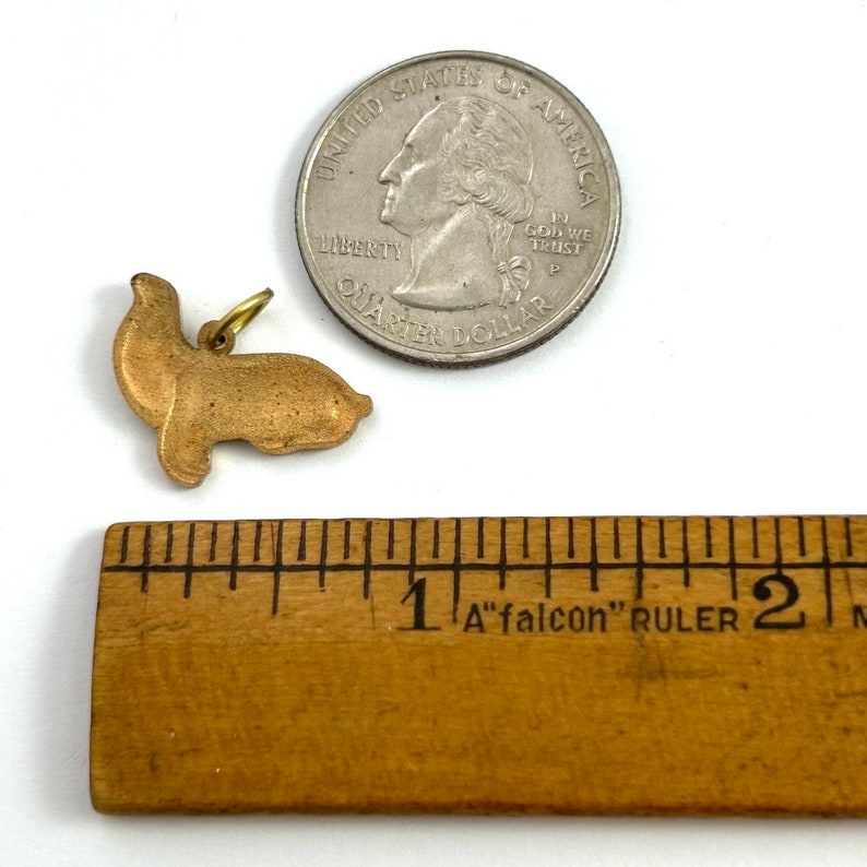 May include: A gold-colored metal charm shaped like a seal. The charm is about 1 inch long and is next to a US quarter for size comparison.