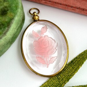 May include: A delicate pink rose carved into a clear glass oval pendant, set in a gold-toned metal frame. The pendant hangs from a gold-toned chain.
