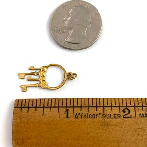 Op de afbeelding: Gouden sleutelbedel met vijf sleutels bevestigd aan een ronde ring. De bedel is ongeveer 2,5 cm in diameter en wordt getoond naast een liniaal ter vergelijking van de grootte.