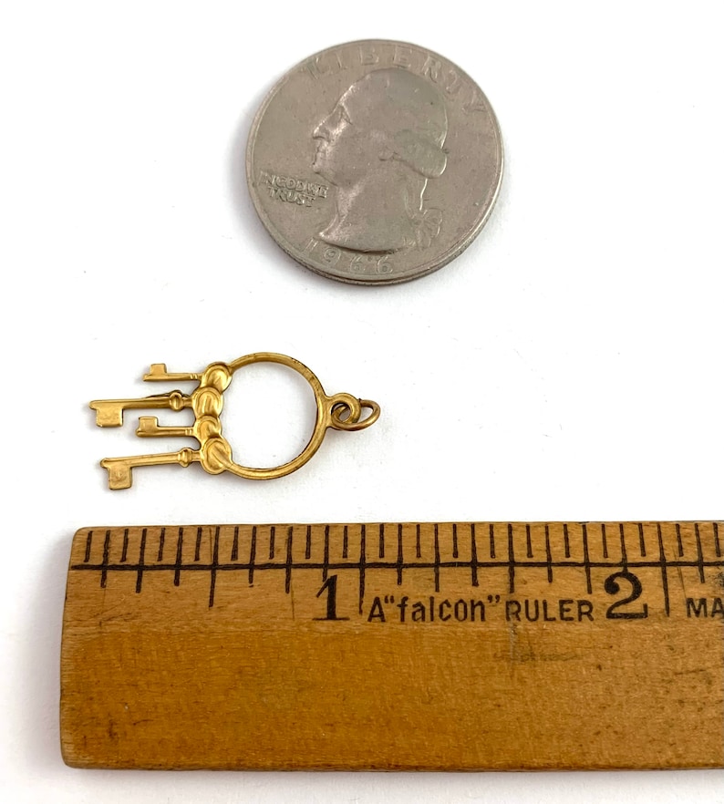 Op de afbeelding: Gouden sleutelbedel met vijf sleutels bevestigd aan een ronde ring. De bedel is ongeveer 2,5 cm in diameter en wordt getoond naast een liniaal ter vergelijking van de grootte.