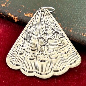 Puede incluir: Colgante plateado con forma de abanico, con intrincados dise&ntilde;os grabados. El dise&ntilde;o incluye l&iacute;neas curvas y patrones decorativos. Un peque&ntilde;o anillo plateado est&aacute; unido en la parte superior. El colgante se muestra sobre una superficie de terciopelo rojo.