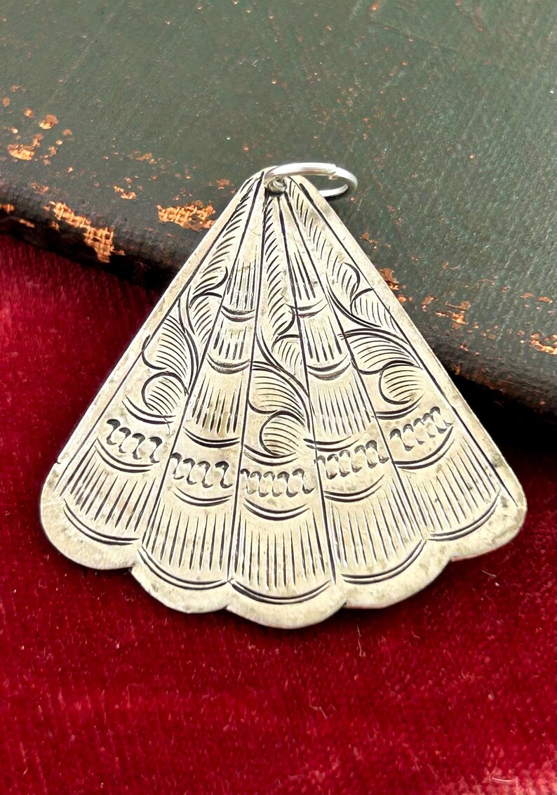 Puede incluir: Colgante plateado con forma de abanico, con intrincados dise&ntilde;os grabados. El dise&ntilde;o incluye l&iacute;neas curvas y patrones decorativos. Un peque&ntilde;o anillo plateado est&aacute; unido en la parte superior. El colgante se muestra sobre una superficie de terciopelo rojo.