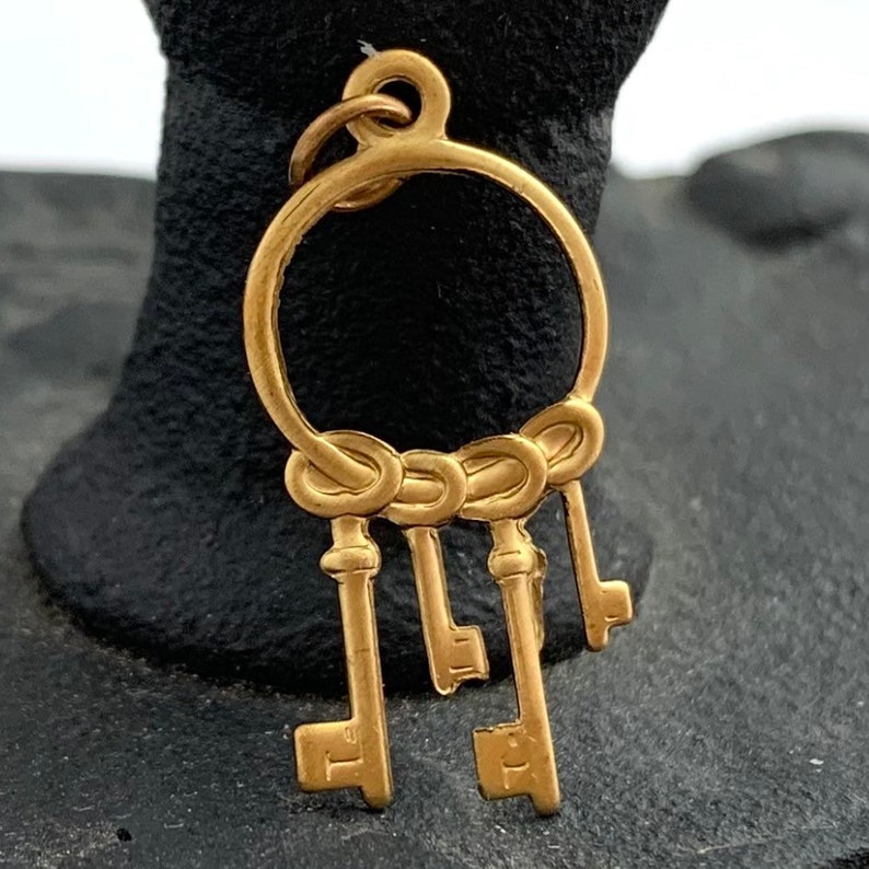 Op de afbeelding: Gouden bedel met een cirkel waaraan vijf sleutels hangen. De sleutels zijn met een reeks knopen aan de cirkel verbonden.