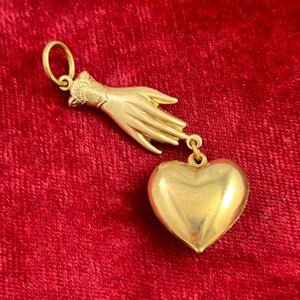 May include: A gold charm pendant featuring a hand reaching out to a heart. The hand and heart are both made of gold and have a shiny finish.