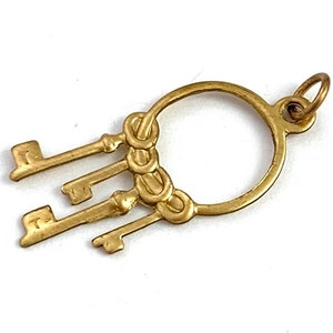 Op de afbeelding: Gouden hanger met een cirkel waaraan vier sleutels hangen. De sleutels zijn met een decoratief knoopje aan de cirkel bevestigd.