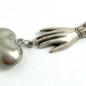 Puede incluir: Collar con dije de plata con un colgante en forma de coraz&oacute;n y una mano extendida. La mano est&aacute; detallada con un brazalete en la mu&ntilde;eca.