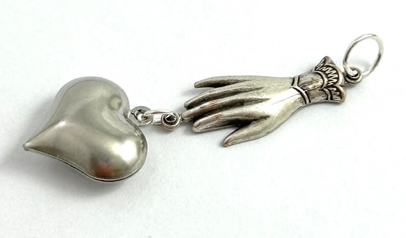 Puede incluir: Collar con dije de plata con un colgante en forma de coraz&oacute;n y una mano extendida. La mano est&aacute; detallada con un brazalete en la mu&ntilde;eca.