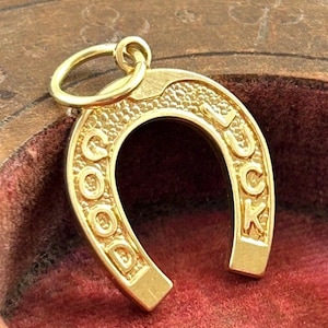 May include: A gold-colored horseshoe charm with the words "GOOD LUCK" embossed on it. The charm has a textured surface and a small loop at the top for attaching to a necklace or bracelet. The charm is displayed against a red background.
