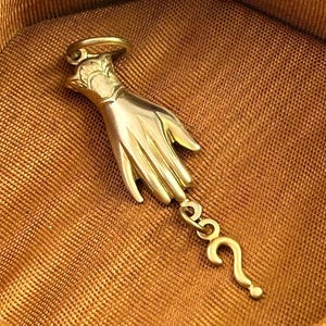K&ouml;nnte beinhalten: Ein goldfarbener Anh&auml;nger mit einer detaillierten Hand und einem Fragezeichen-Anh&auml;nger, der daran h&auml;ngt. Der Anh&auml;nger befindet sich auf einem braunen Stoffhintergrund.