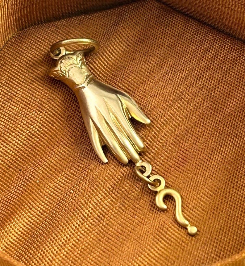 K&ouml;nnte beinhalten: Ein goldfarbener Anh&auml;nger mit einer detaillierten Hand und einem Fragezeichen-Anh&auml;nger, der daran h&auml;ngt. Der Anh&auml;nger befindet sich auf einem braunen Stoffhintergrund.