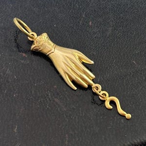 K&ouml;nnte beinhalten: Ein goldener Anh&auml;nger in Form einer Hand mit einem Fragezeichen, das am Handgelenk baumelt.