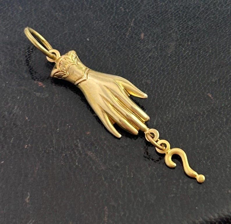 K&ouml;nnte beinhalten: Ein goldener Anh&auml;nger in Form einer Hand mit einem Fragezeichen, das am Handgelenk baumelt.