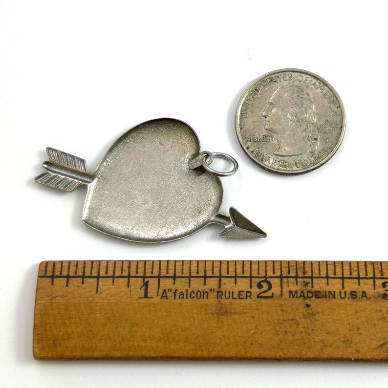 Może przedstawiać: Srebrny charm w kształcie serca z przechodzącą przez niego strzałą. Charm ma około 2,5 cm średnicy i ma pętelkę do przymocowania do naszyjnika lub bransoletki.