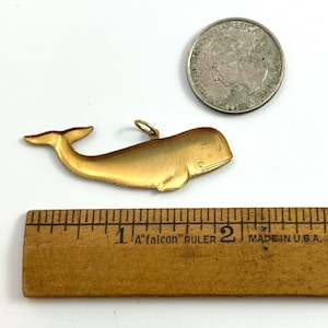 May include: A gold-coloured whale charm pendant, approximately 2.5 cm long, with a loop for attaching to a chain or bracelet. The charm is shown next to a ruler and a US quarter for size reference.