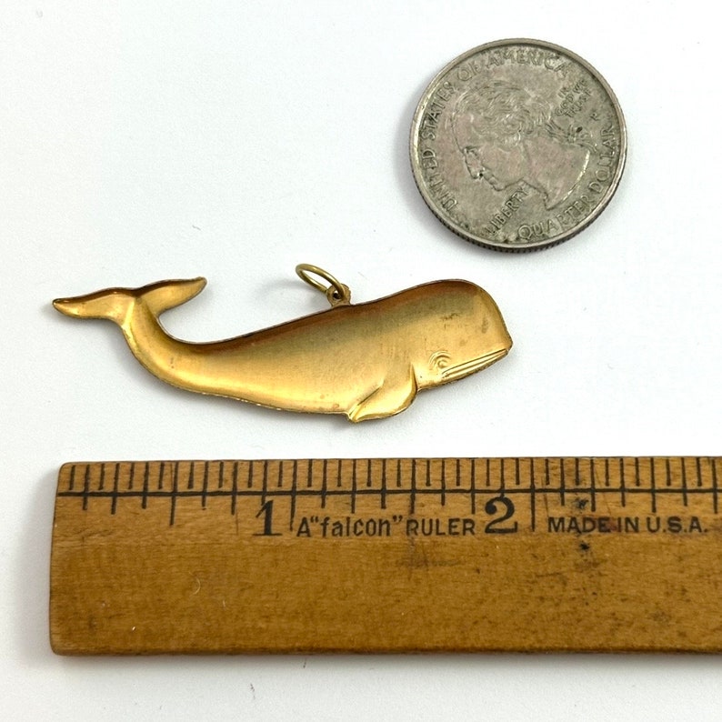 May include: A gold-coloured whale charm pendant, approximately 2.5 cm long, with a loop for attaching to a chain or bracelet. The charm is shown next to a ruler and a US quarter for size reference.