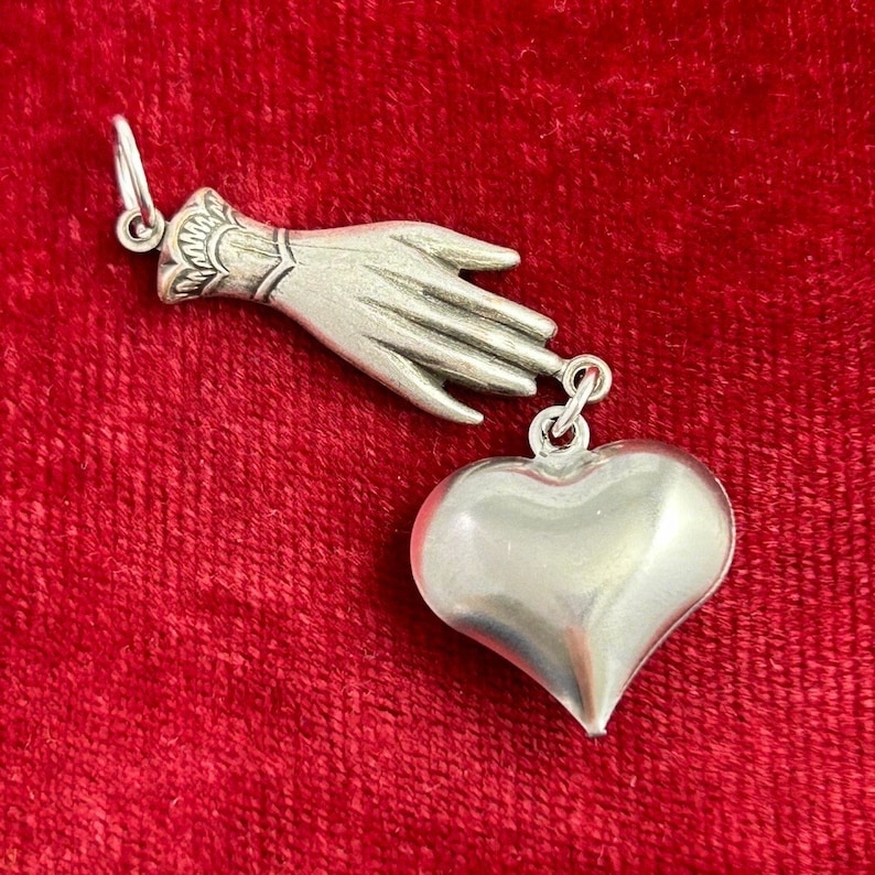 Puede incluir: Un charm de plata con una mano que alcanza un coraz&oacute;n. La mano est&aacute; detallada con un dise&ntilde;o texturizado y el coraz&oacute;n es liso y redondeado. El charm es perfecto para agregar un toque de sentimentalismo a cualquier pieza de joyer&iacute;a.