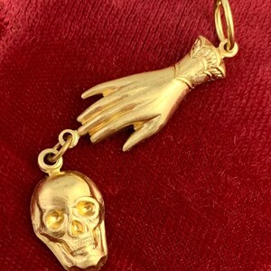 Op de afbeelding: Een gouden ketting met een bedel in de vorm van een hand die naar een schedel reikt.