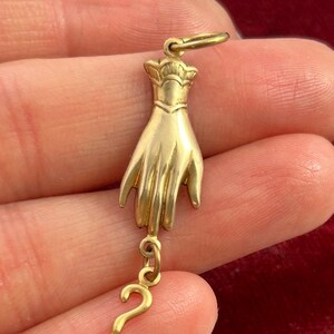 K&ouml;nnte beinhalten: Ein goldfarbener Anh&auml;nger in Form einer Hand mit einem Fragezeichen, das am Handgelenk h&auml;ngt. Der Anh&auml;nger ist mit einer strukturierten Oberfl&auml;che und einem kronenartigen Design an der Oberseite verziert.