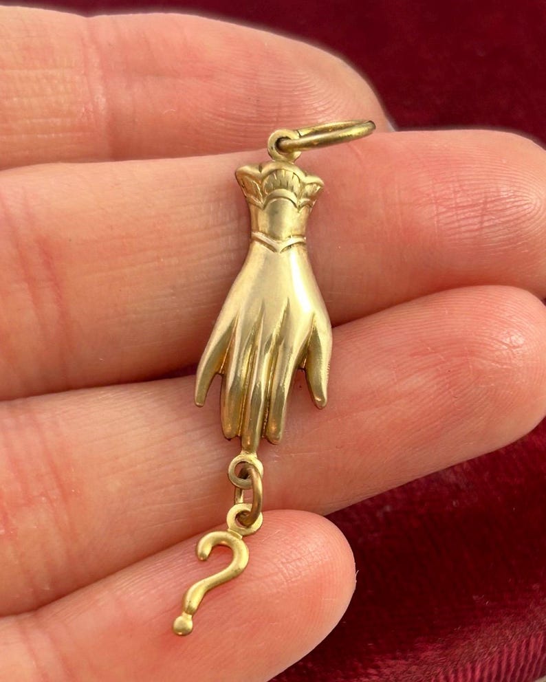 K&ouml;nnte beinhalten: Ein goldfarbener Anh&auml;nger in Form einer Hand mit einem Fragezeichen, das am Handgelenk h&auml;ngt. Der Anh&auml;nger ist mit einer strukturierten Oberfl&auml;che und einem kronenartigen Design an der Oberseite verziert.