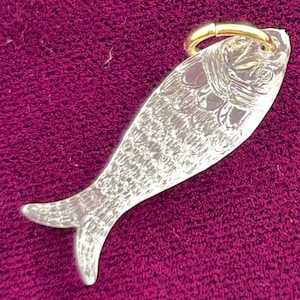 May include: A silver-colored fish-shaped pendant with etched details, including scales and a visible eye. The pendant has a gold-colored ring at the top for attachment. The fish is displayed against a deep purple textured background.