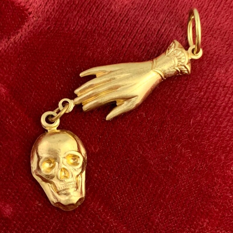 Op de afbeelding: Een gouden hanger met een hand die naar een schedel reikt. De hand is versierd met een manchet en de schedel heeft een getextureerd oppervlak.