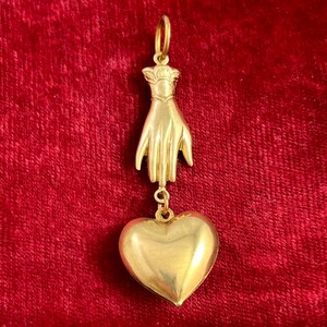 May include: A gold pendant featuring a hand holding a heart. The pendant is made of solid gold and has a detailed design.
