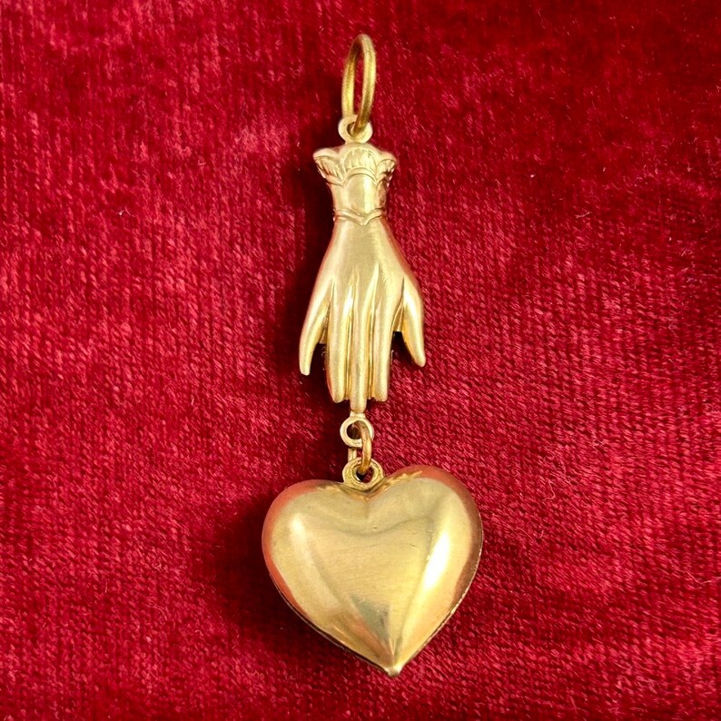 May include: A gold pendant featuring a hand holding a heart. The pendant is made of solid gold and has a detailed design.