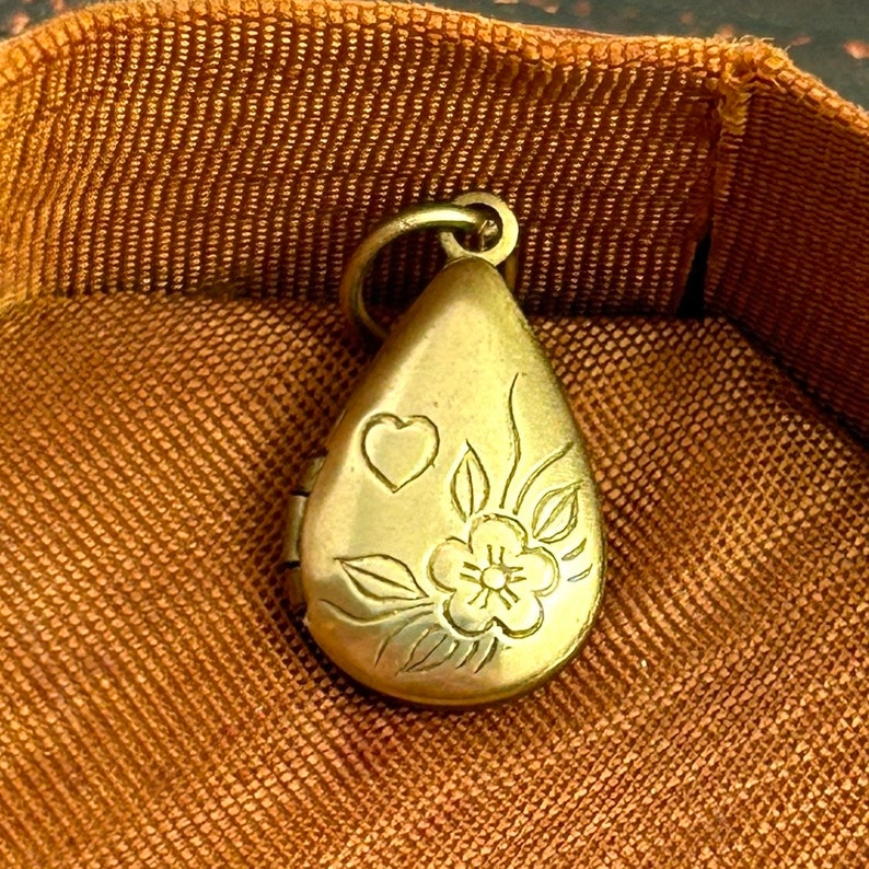 May include: A gold-toned teardrop-shaped locket with a floral design and a small heart engraved on the front.
