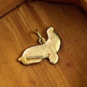 May include: A gold-colored charm in the shape of a seal. The seal is lying on its belly with its head raised and looking to the right. The charm has a loop at the top for attaching to a necklace or bracelet.