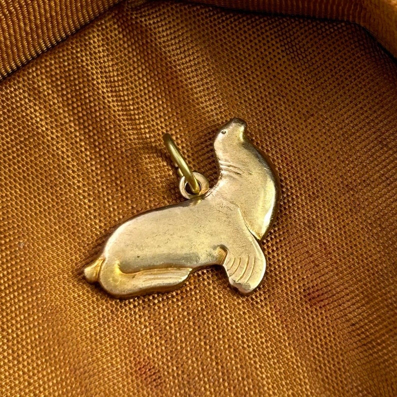 May include: A gold-colored charm in the shape of a seal. The seal is lying on its belly with its head raised and looking to the right. The charm has a loop at the top for attaching to a necklace or bracelet.