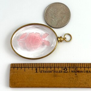 May include: A gold-toned oval locket with a clear glass front featuring a pink rose carved into the glass. The locket has a gold-toned chain attached to it.