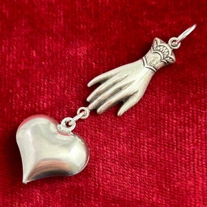 Puede incluir: Un collar con un dije de plata que representa una mano que se extiende hacia un coraz&oacute;n. La mano est&aacute; detallada con un brazalete y el coraz&oacute;n es liso y redondeado.