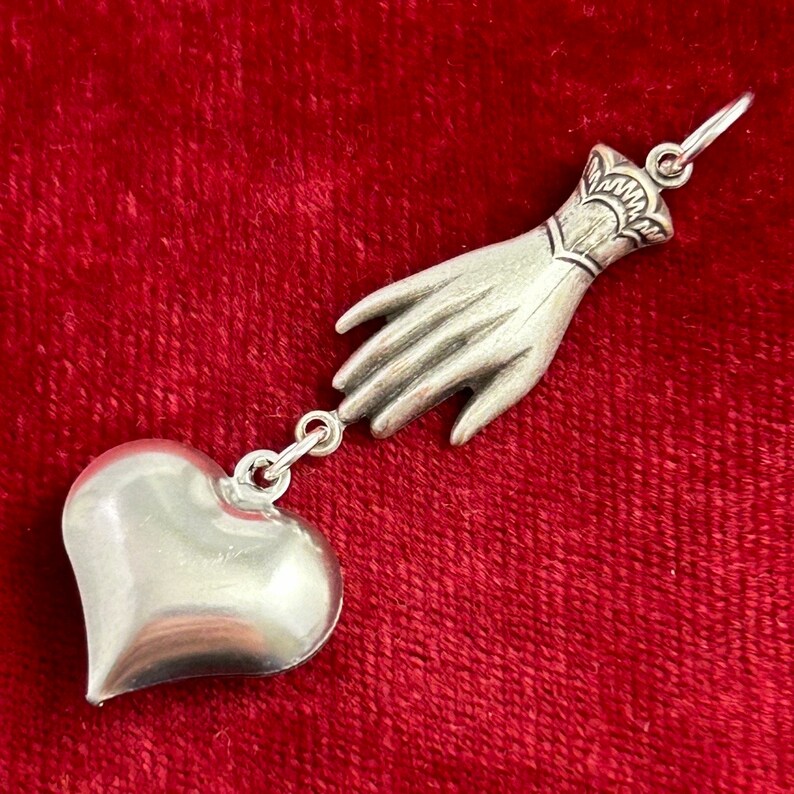 Puede incluir: Un collar con un dije de plata que representa una mano que se extiende hacia un coraz&oacute;n. La mano est&aacute; detallada con un brazalete y el coraz&oacute;n es liso y redondeado.
