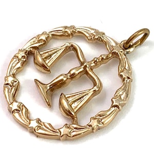 May include: Gold-toned pendant charm featuring a stylized depiction of the scales of justice, surrounded by a circular frame with star-shaped accents.