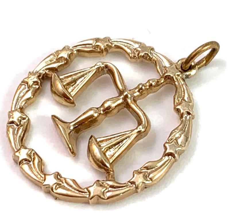 May include: Gold-toned pendant charm featuring a stylized depiction of the scales of justice, surrounded by a circular frame with star-shaped accents.