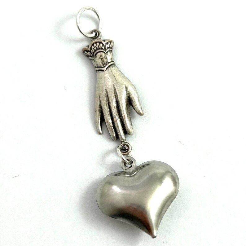 Puede incluir: Encanto de colgante de plata con una mano sosteniendo un coraz&oacute;n. La mano est&aacute; detallada con un brazalete y el coraz&oacute;n es liso y redondeado.