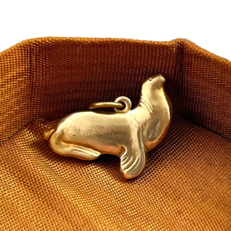 May include: A gold-colored charm in the shape of a seal. The charm is lying on a brown fabric surface.