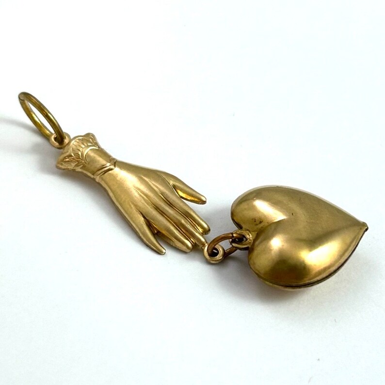 May include: A gold-toned pendant featuring a hand reaching towards a heart-shaped locket. The hand is detailed with a cuff and the heart is smooth and shiny.